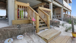 Un escalier en bois mène à un bâtiment doté d’un balcon, lequel est entouré de sable et de quelques plantes.