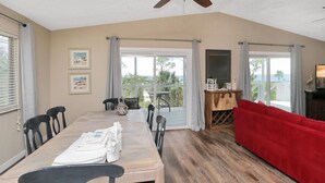 A spacious room with a dining table, chairs, a red sofa, a ceiling fan, and large windows offering a view of the outdoors. - Hideaway Bay (Port St. Joe)