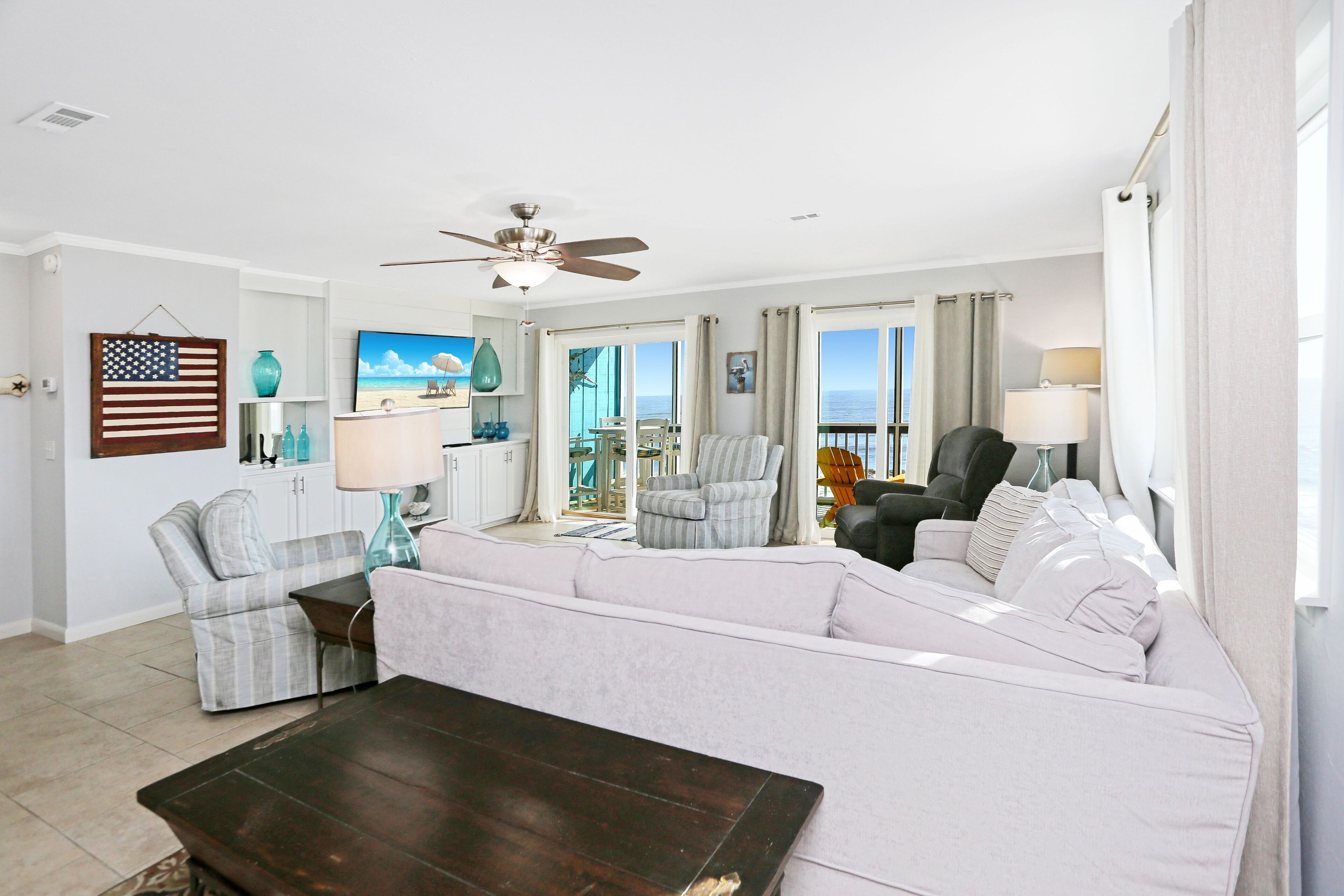 A spacious living room with a large white sofa, a dark wooden coffee table, and a ceiling fan. There is a view of the ocean through large windows, and a small table with a lamp is visible.