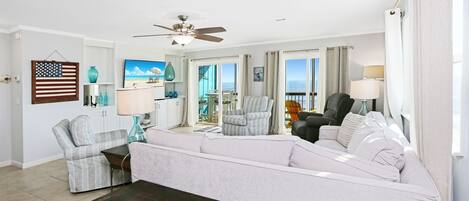 A spacious living room with a large white sofa, a dark wooden coffee table, and a ceiling fan. There is a view of the ocean through large windows, and a small table with a lamp is visible.