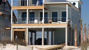 Une maison de deux étages située en bord de mer, dotée d’un balcon, d’une terrasse couverte au rez-de-chaussée et d’une plage de sable au premier plan.