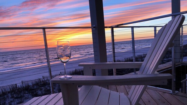 Une terrasse en bois, un verre de vin et une chaise d’où l’on peut admirer le coucher de soleil sur la plage.