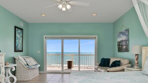 A bedroom with a bed, a chair, a sofa, a ceiling fan, and a balcony with a view of the ocean.