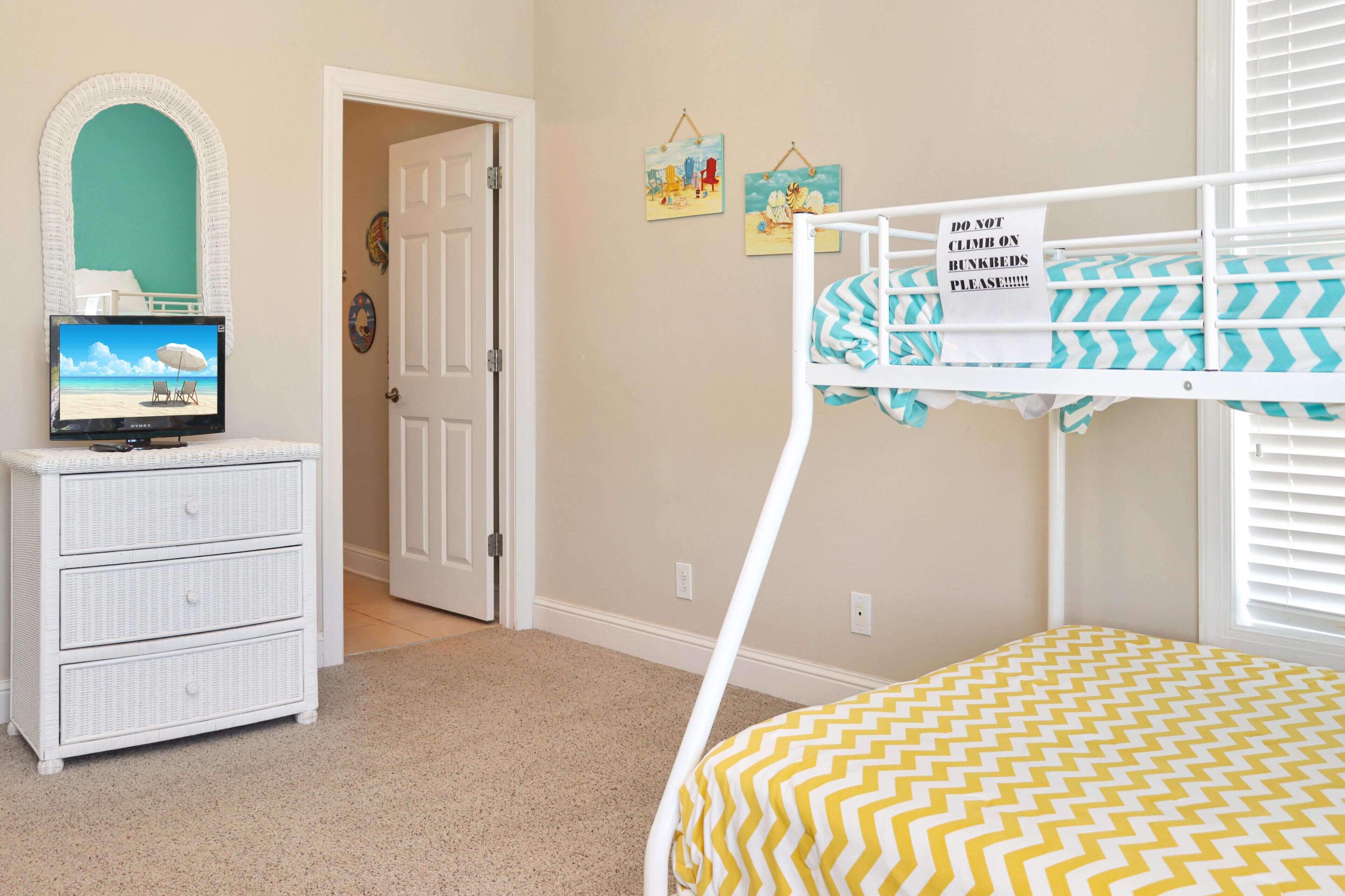 A child's bedroom with bunk beds, a dresser, a television, and a door leading to another room.