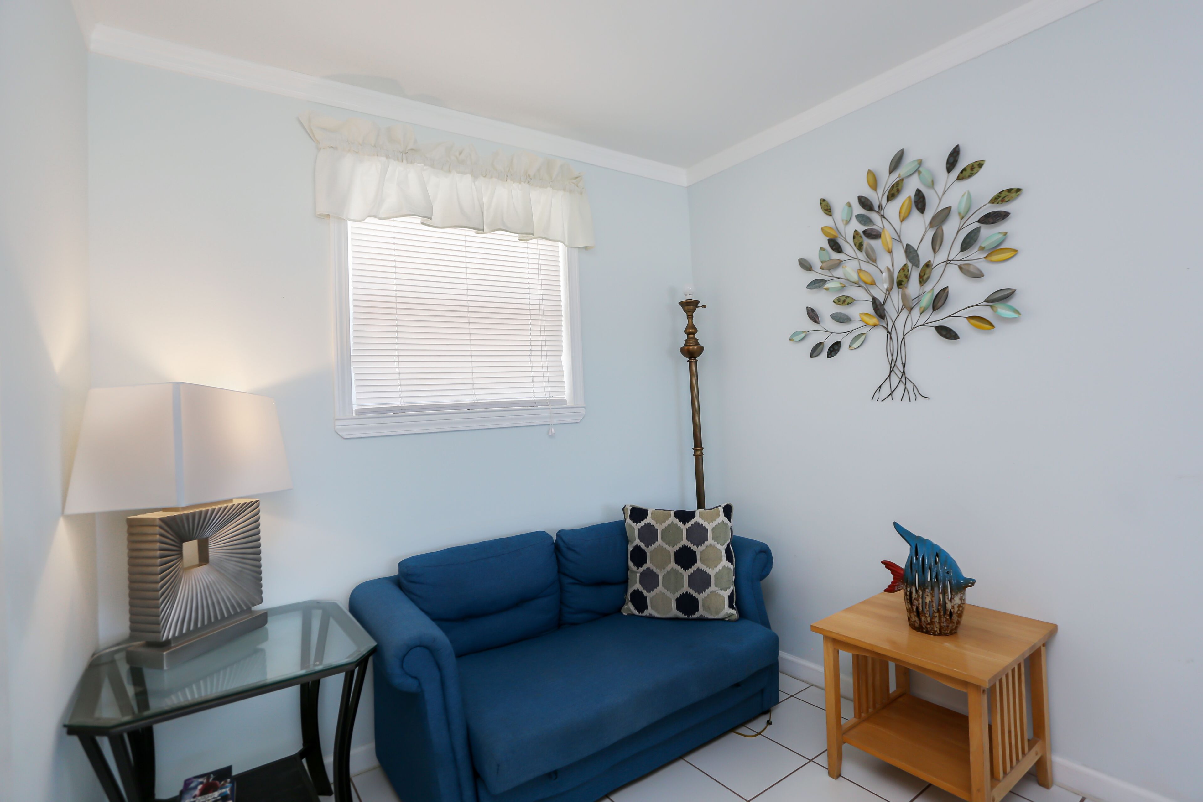 A living room with a blue sofa, a glass coffee table, a wooden side table, a wall-mounted lamp, a window with blinds, and a decorative tree artwork.