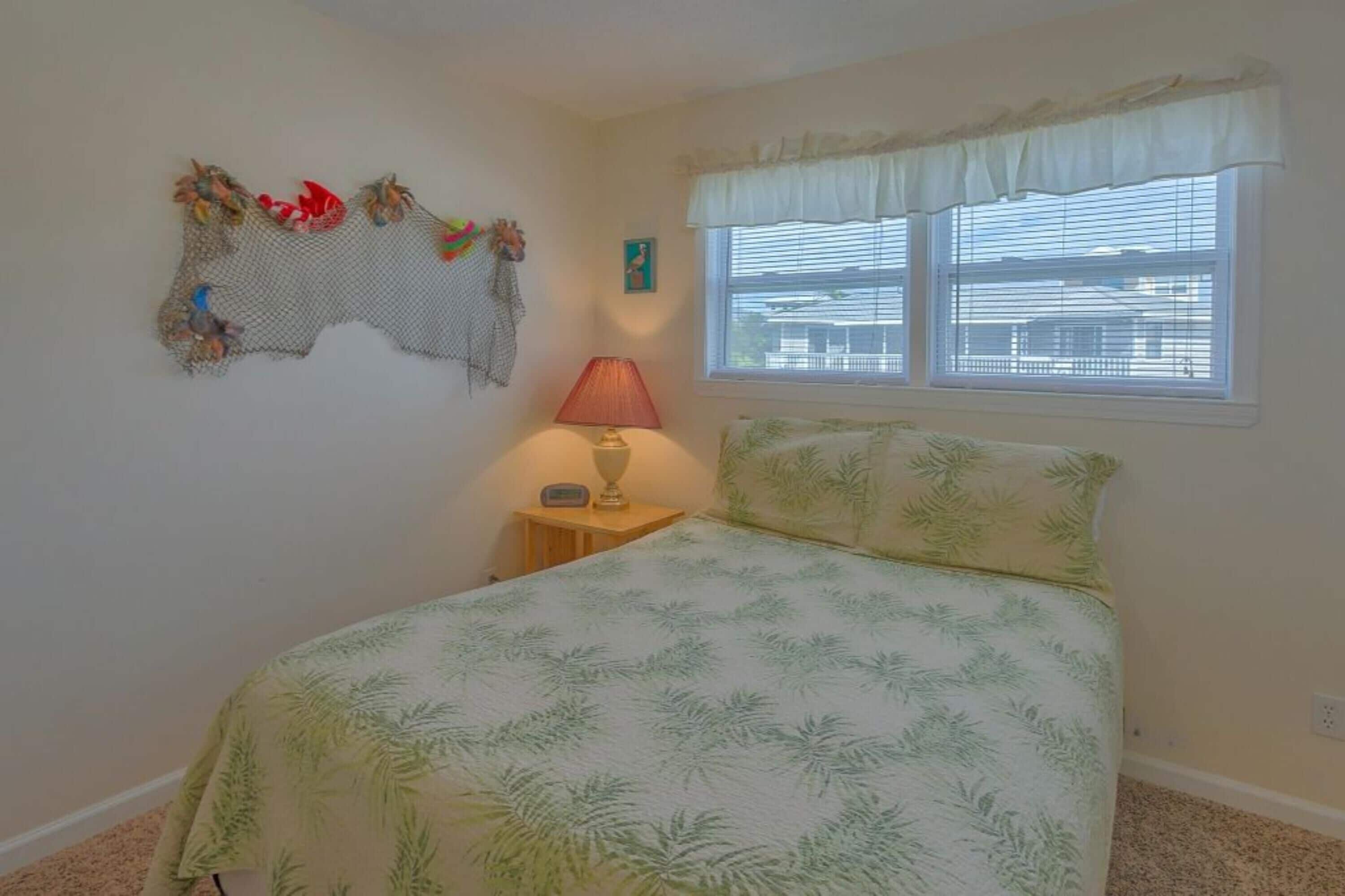 A bedroom with a bed, a nightstand, a lamp, a window with blinds, and a wall decoration.