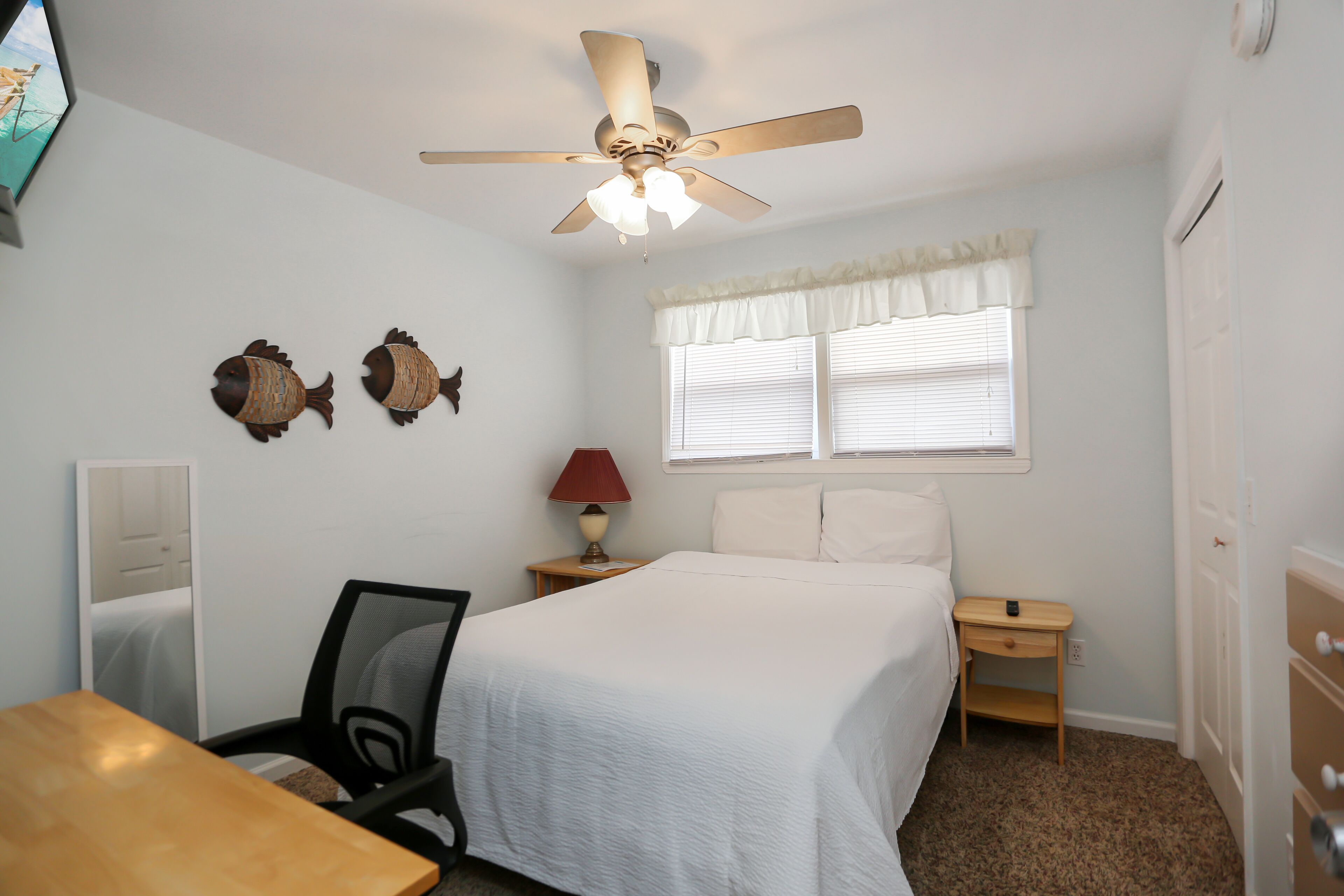 A bedroom with a bed, a desk, a chair, a ceiling fan, a window with blinds, and a nightstand with a lamp.
