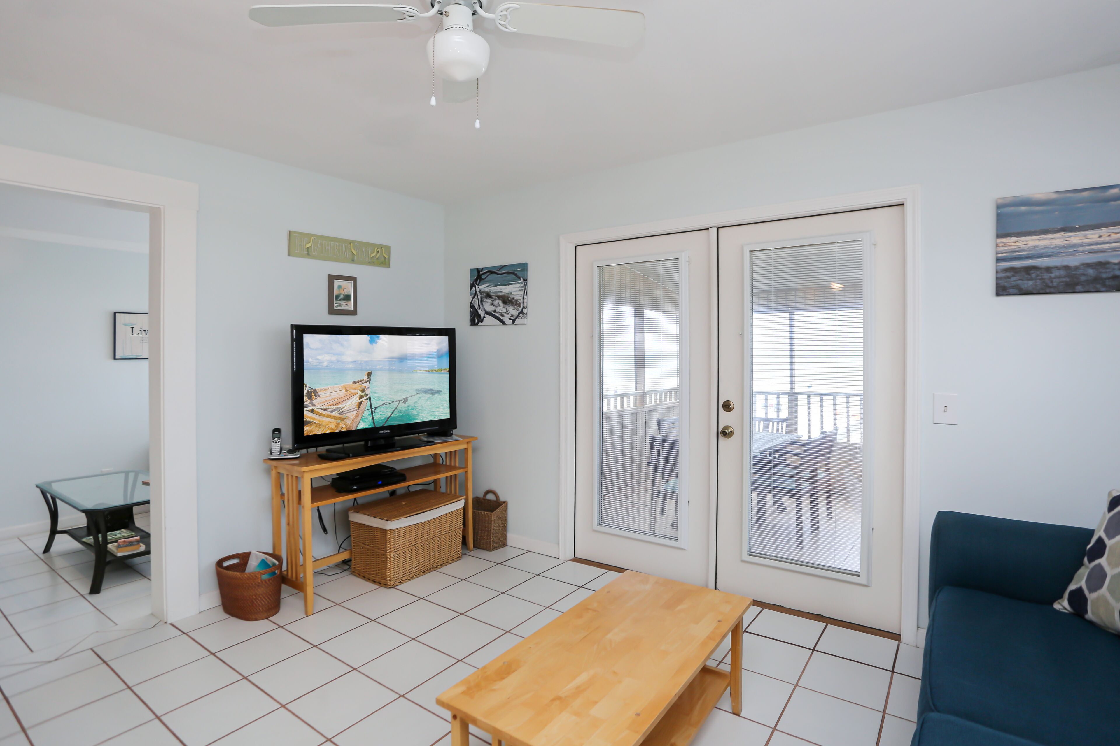 Uma sala de estar com televisão sobre um suporte de madeira, um sofá azul, uma mesa de madeira e uma porta para varanda com persianas.
