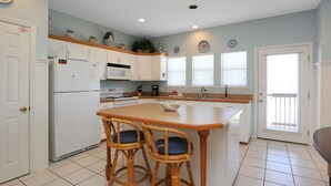 A kitchen with white cabinets, a central island with two bar stools, a refrigerator, a microwave, and a coffee maker. - The Shore House (Port St. Joe)