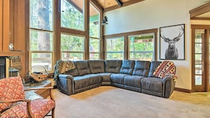 A spacious living room with a large sectional sofa, a fireplace, and large windows offering a view of the outdoors. - Cabin < Half-Mi from Pine Mountain Lake Marina! (Groveland)