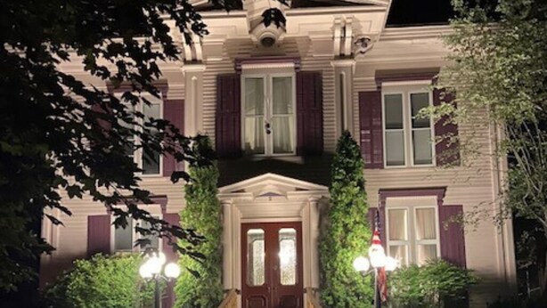 Ein zweistöckiges Gebäude mit einem zentralen Eingang, flankiert von zwei Fenstern mit Fensterläden, und einer kleinen Veranda mit Stufen zum Eingang.