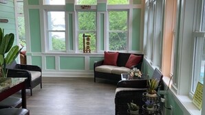 Ein gemütliches Zimmer mit großem Fenster, einer Couch mit Kissen, einem kleinen Tisch mit Pflanzen und einem Sessel.