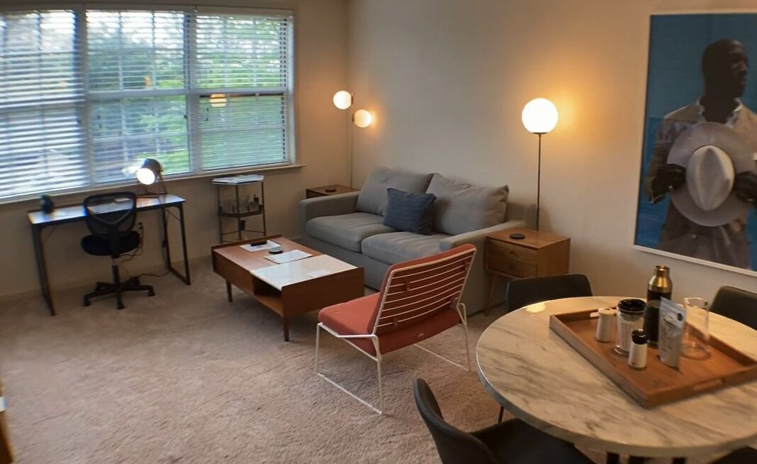 Ett mysigt vardagsrum med en soffa, ett matbord med ett bricka på, ett skrivbord med en lampa och ett stort fönster med persienner.