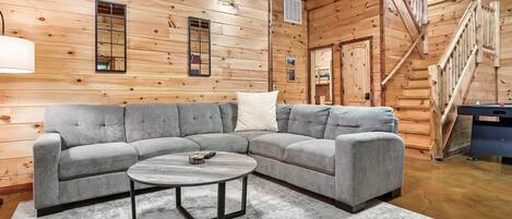 A log cabin interior with a gray sectional sofa, a round coffee table, wooden walls, a staircase, and a small window.