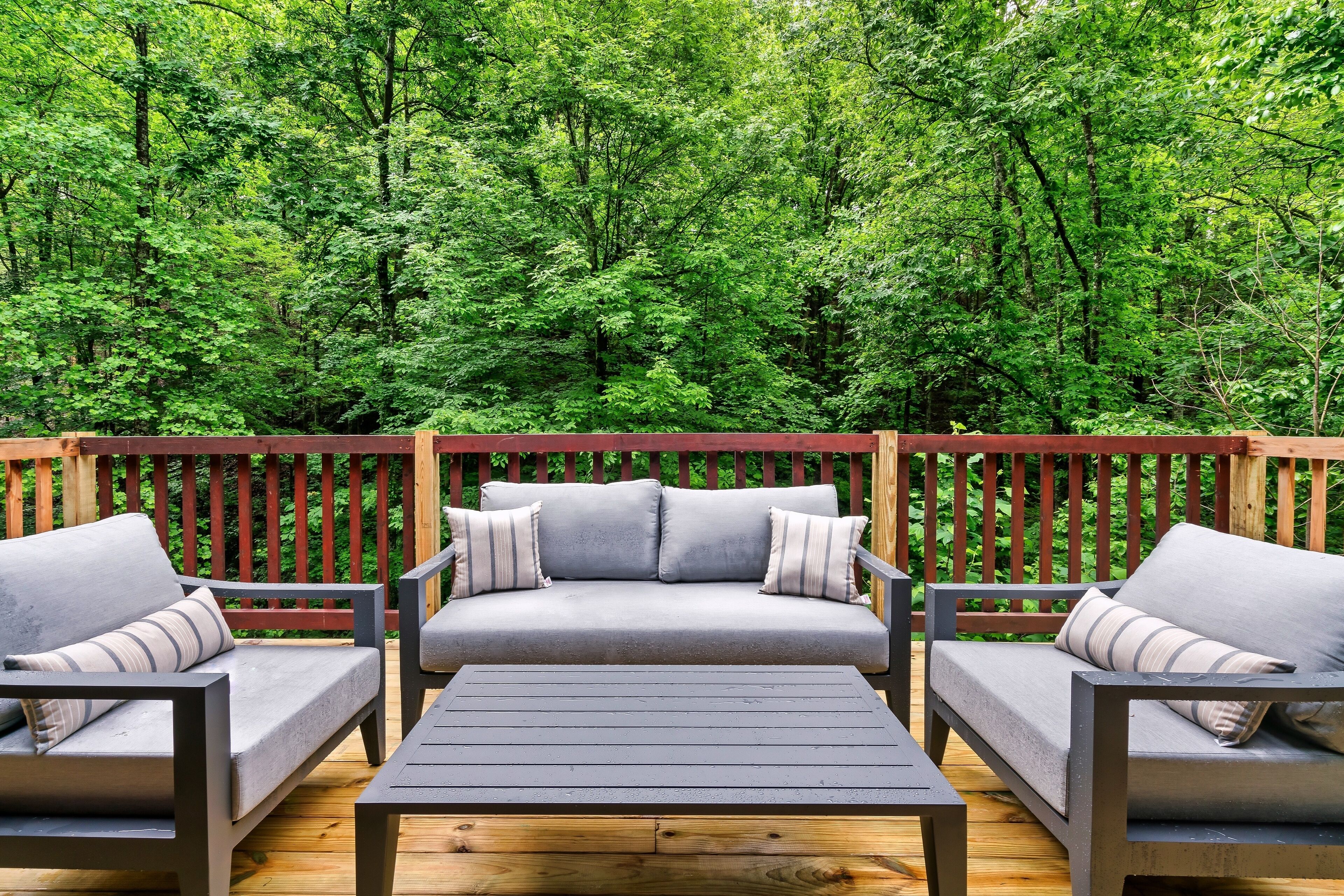 An outdoor deck with a gray sofa and two armchairs, a wooden table, and a wooden railing.