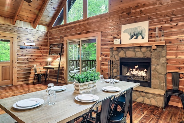 A wooden cabin interior with a dining table set for four, a fireplace, and a large window overlooking greenery.