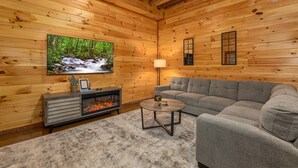 Ein gemütliches Blockhaus-Interieur mit Kamin, einem grauen Sofa, einem runden Couchtisch und einem an der Wand befestigten Fernseher, der eine Waldszene zeigt.