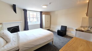 A hotel room with a bed, a chair, a small table, a mirror, a window with curtains, a radiator, and a wall-mounted light fixture.