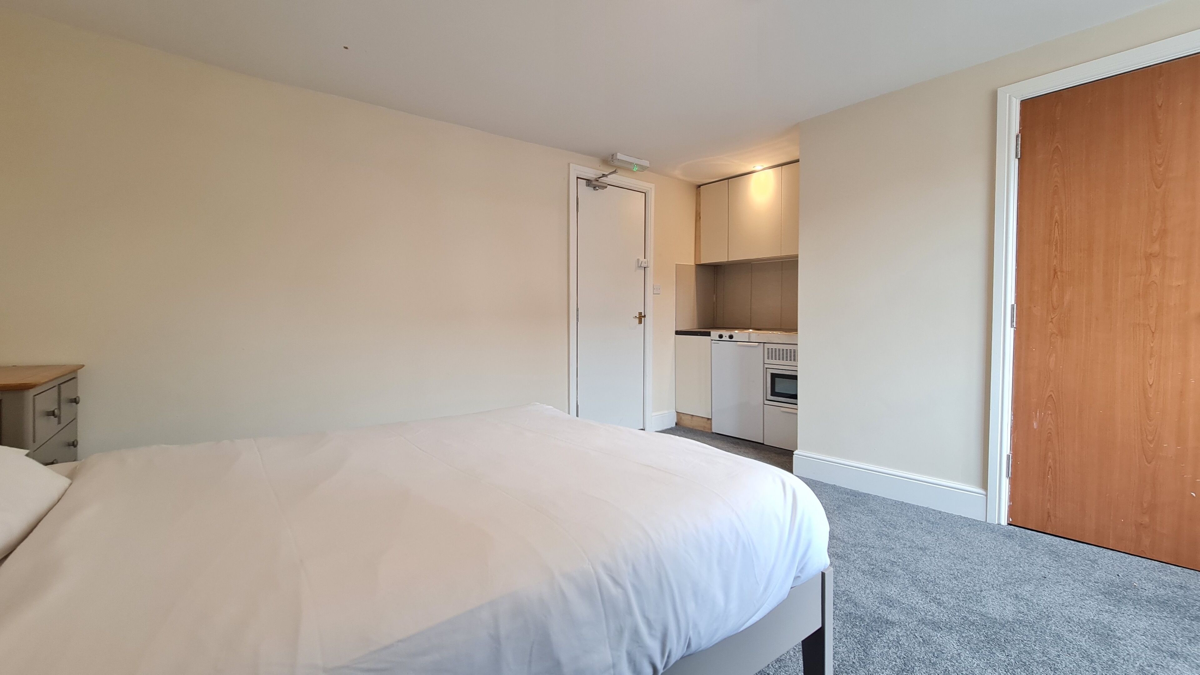 A bedroom with a bed, a wooden door, and a kitchenette in the background.