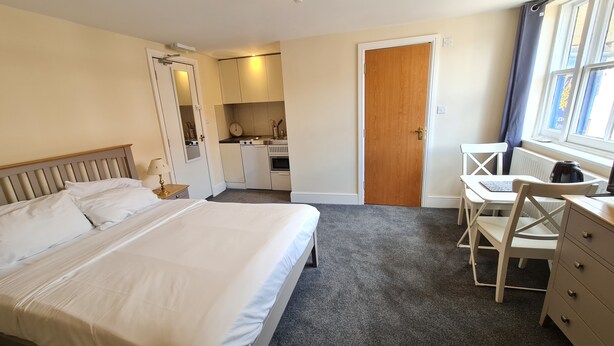 Une chambre à coucher avec un lit, un bureau, une chaise, une commode et, au fond, un coin cuisine.