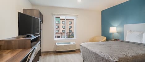 A hotel room with a bed, a desk with a TV, a chair, a window, and a lamp.