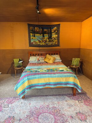 A bedroom with a wooden bed, colorful bedding, a tapestry, and two chairs.