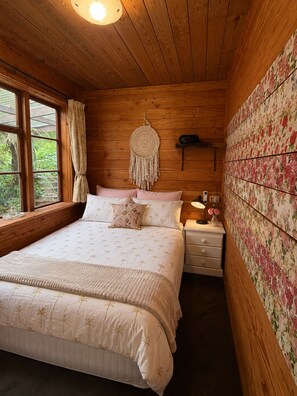 A wooden room with a bed, a nightstand, a wall with floral wallpaper, and a window with curtains.