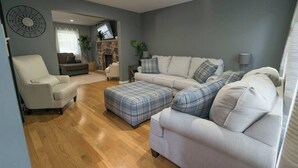 A living room with a grey wall, a white sofa, a grey armchair, and a plaid ottoman. - Lake Adjacent Home w/ Hot Tub, Game Room, Boats, Fenced Yard (East Stroudsburg)