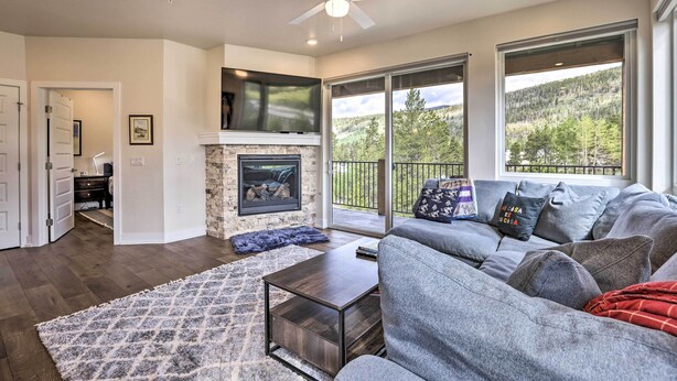 A spacious living room with a fireplace, a large gray sofa, and a view of a mountainous landscape through the windows. - Walk to Lifts: Condo w/ Mtn Views in Keystone! (Keystone)