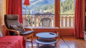 A room with a wooden interior, a red bedspread, a chair, a small table, and a view of mountains and buildings through large windows. - Cosy apartment for 4 people with WIFI, TV and balcony (Basse-nendaz)