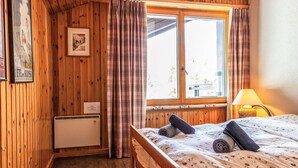 A wooden-paneled room with a bed, a window with curtains, a small table with a lamp, and a wall-mounted air conditioner. - Nice apartment for 8 guests with WIFI, TV and terrace (Basse-nendaz)