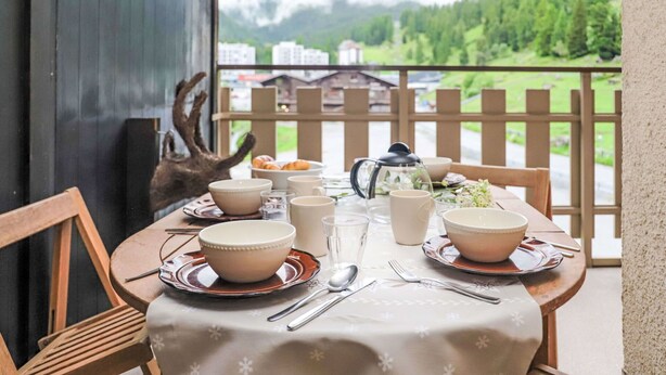 A dining table set with bowls, cups, and a teapot on a balcony with a view of green hills and buildings. - Nice apartment for 4 people with WIFI, TV and balcony (Basse-nendaz)