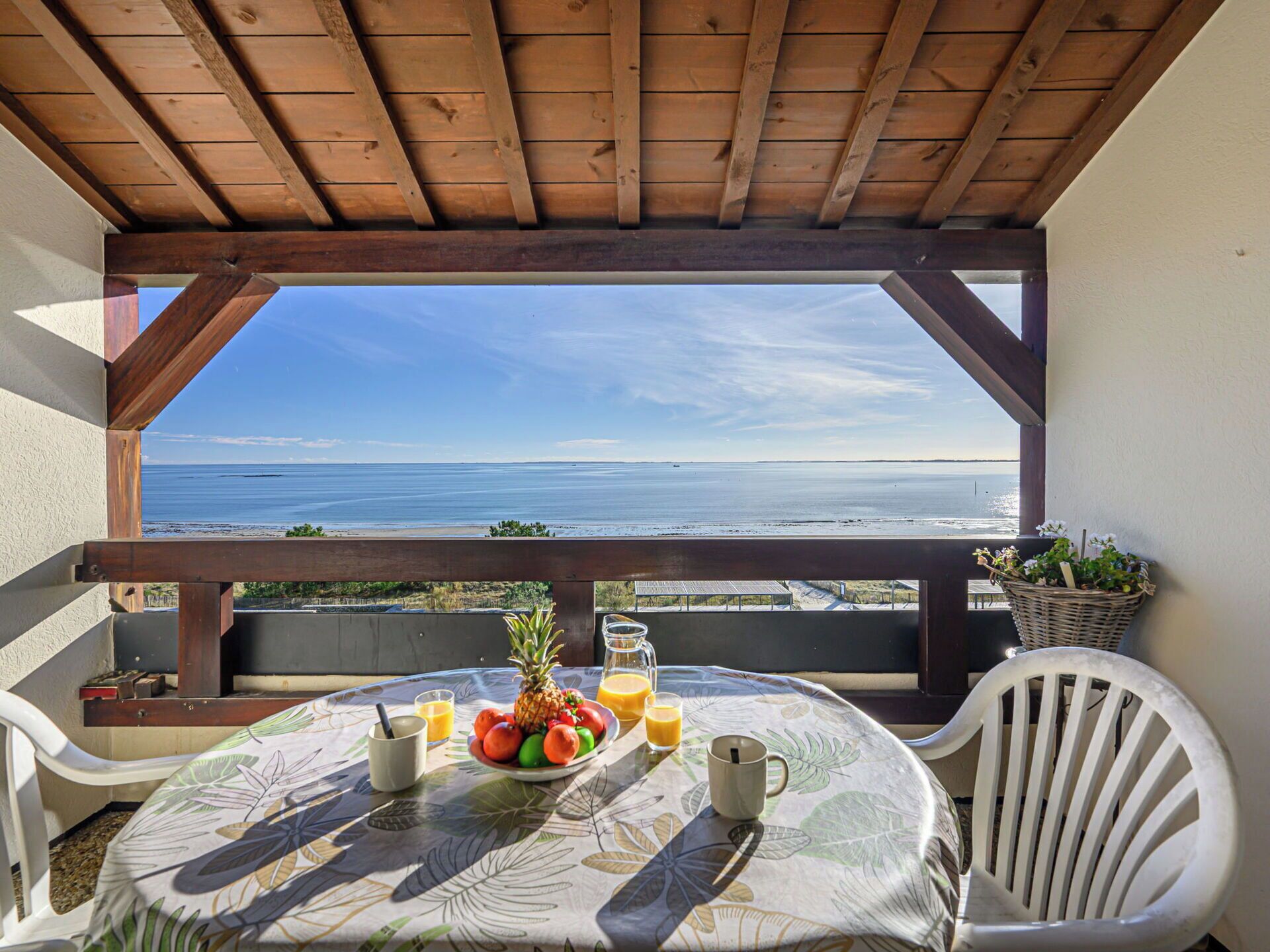 Ein Balkon mit Holzboden, einem Tisch mit Tischdecke aus tropischen Blättern, einer Ananas, Orangen und einem Glas Orangensaft.