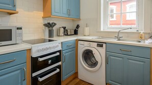 A compact kitchen with blue cabinets, a microwave, an oven, a washing machine, and a window with a view of a red building. - Pebbles Apartment - Heart of Town (North Berwick)