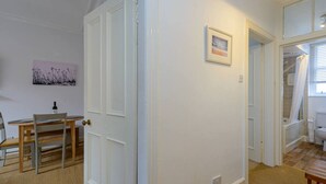 A room with a dining table, chairs, a door, a framed picture, and a shower area. - Pebbles Apartment - Heart of Town (North Berwick)