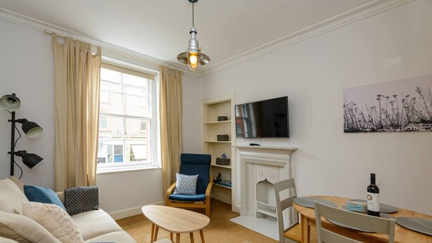 A cozy living room with a sofa, armchair, and a small table. A fireplace is visible, and a TV is mounted on the wall. - Pebbles Apartment - Heart of Town (North Berwick)