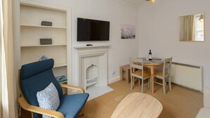 A living room with a fireplace, a television mounted above it, a dining table with chairs, and a blue armchair with a patterned cushion. - Pebbles Apartment - Heart of Town (North Berwick)