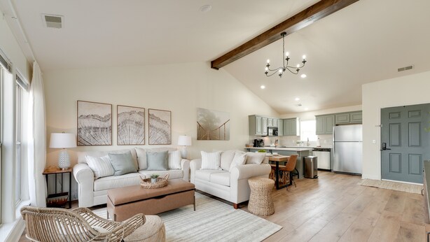 Un espacio habitable amplio con un conjunto de sofás, una mesa de centro y una silla de mimbre. Se ve una cocina con electrodomésticos modernos al fondo.
