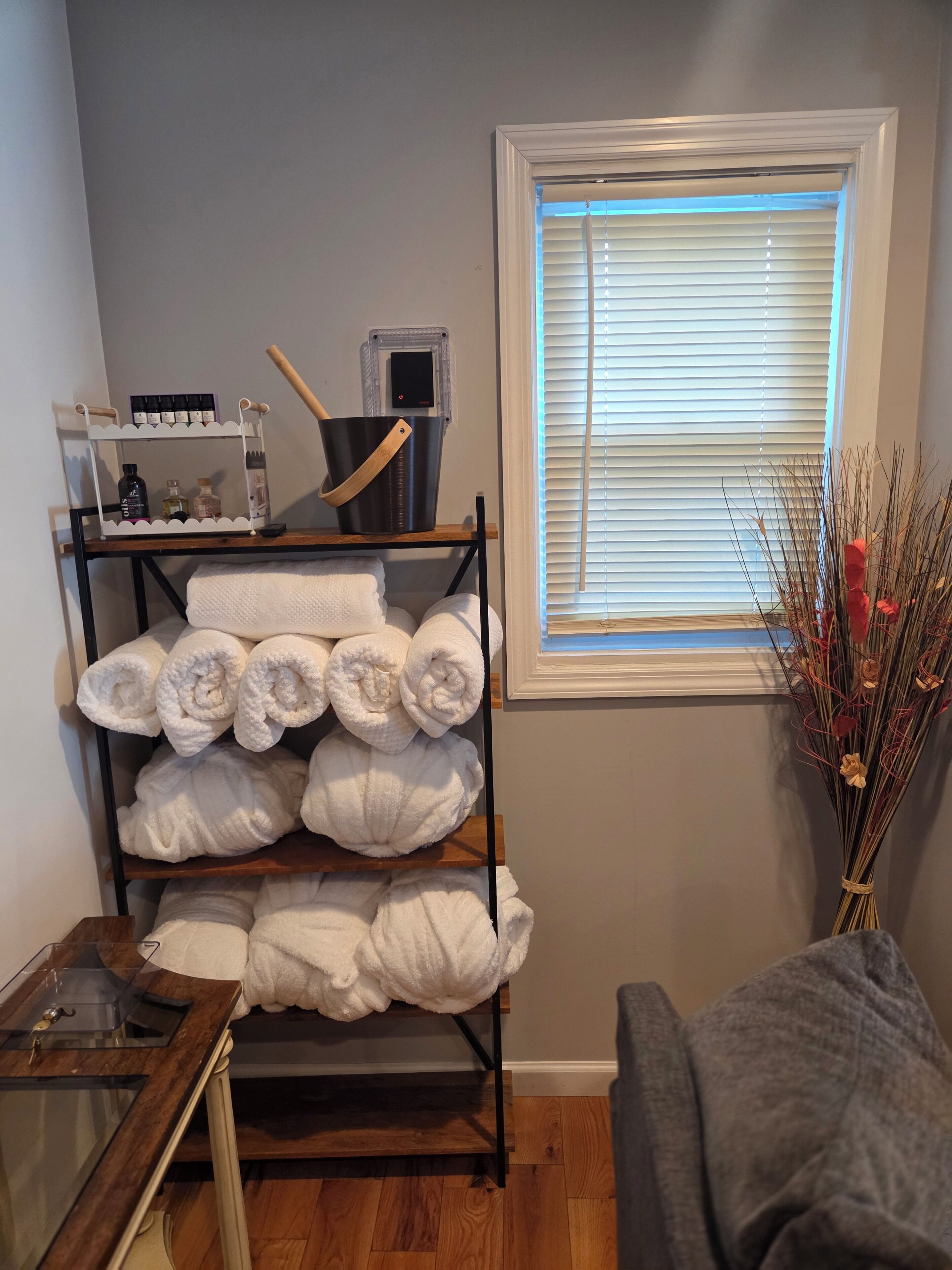 A room with a metal rack holding towels, a small table with a glass container, and a window with blinds.