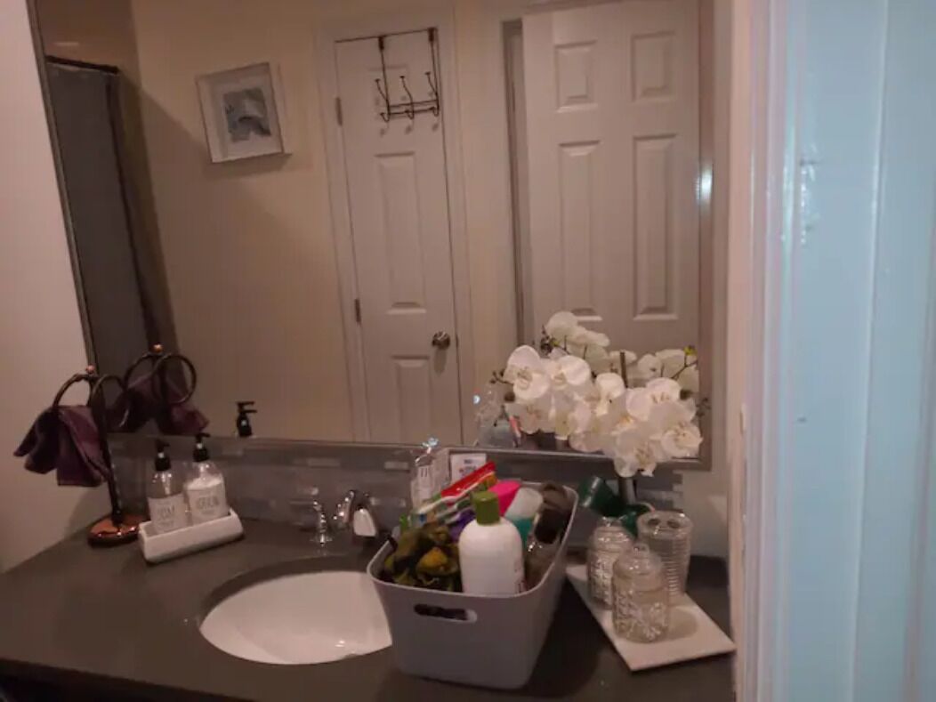 Une salle de bain avec un lavabo blanc, un miroir et un panier rempli d’articles de toilette. On aperçoit des fleurs blanches en arrière-plan et une porte avec un porte-manteau.
