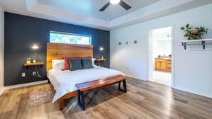 A bedroom with a wooden bed, a bench, a ceiling fan, a nightstand with a lamp, a window, and a view into the kitchen. - 2 Acre FL Oasis with Game Room; Cozy+Quiet - Equestrian and Nature enthusiasts. (Ocala)