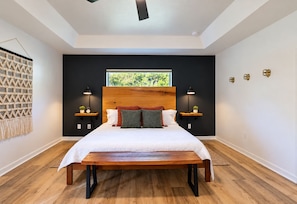 A modern bedroom with a wooden bed, two bedside tables with lamps, a large window, and a macrame wall hanging.