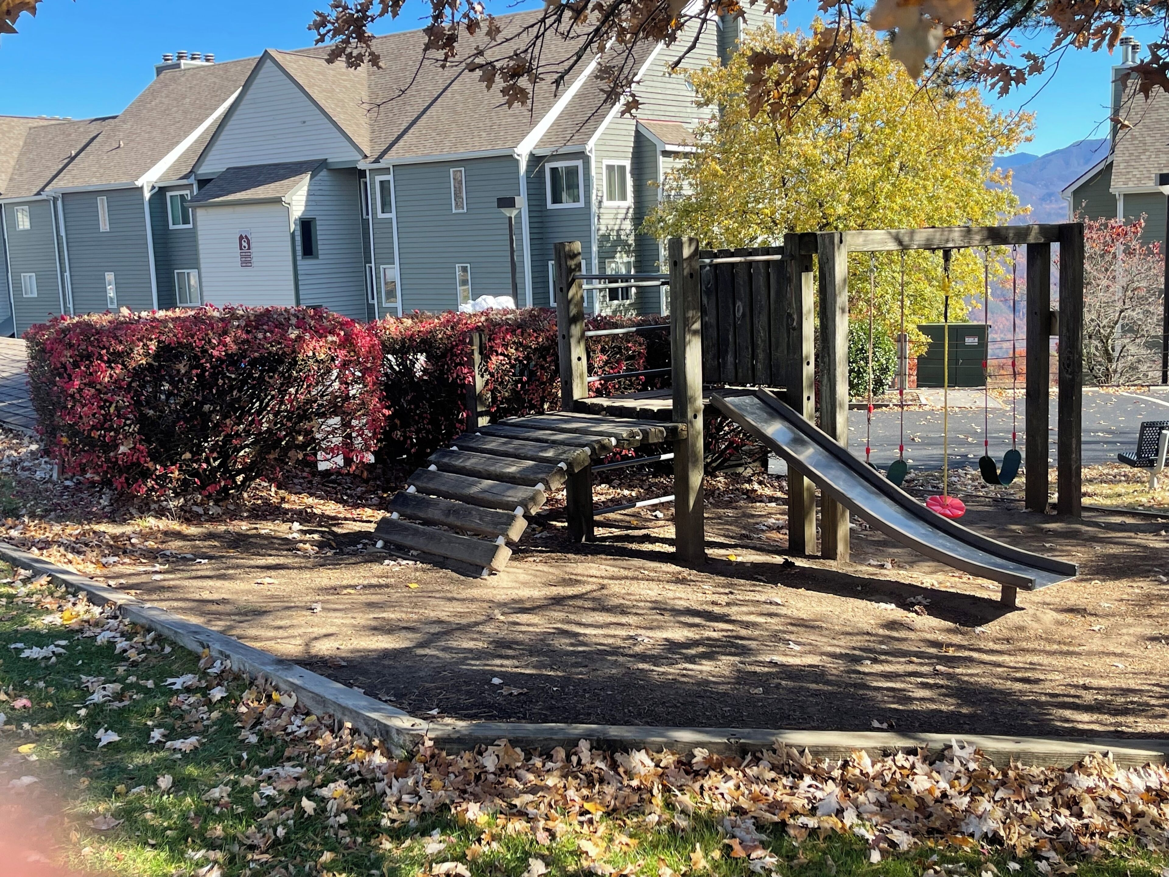 A playground with a slide and steps, surrounded by a hedge and leaf-covered ground.
