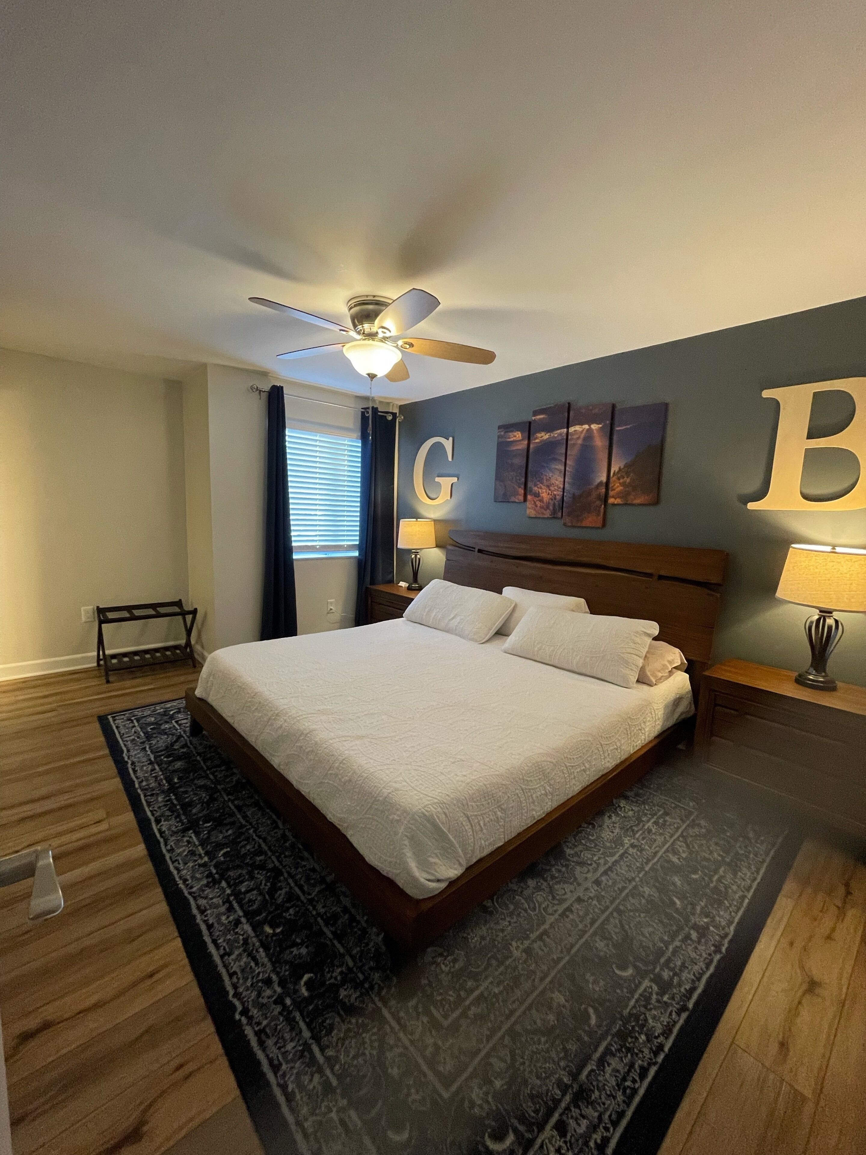 Un dormitorio con una cama grande, un ventilador de techo, una mesita de noche con lámpara, una obra de arte en la pared y una ventana con cortinas.