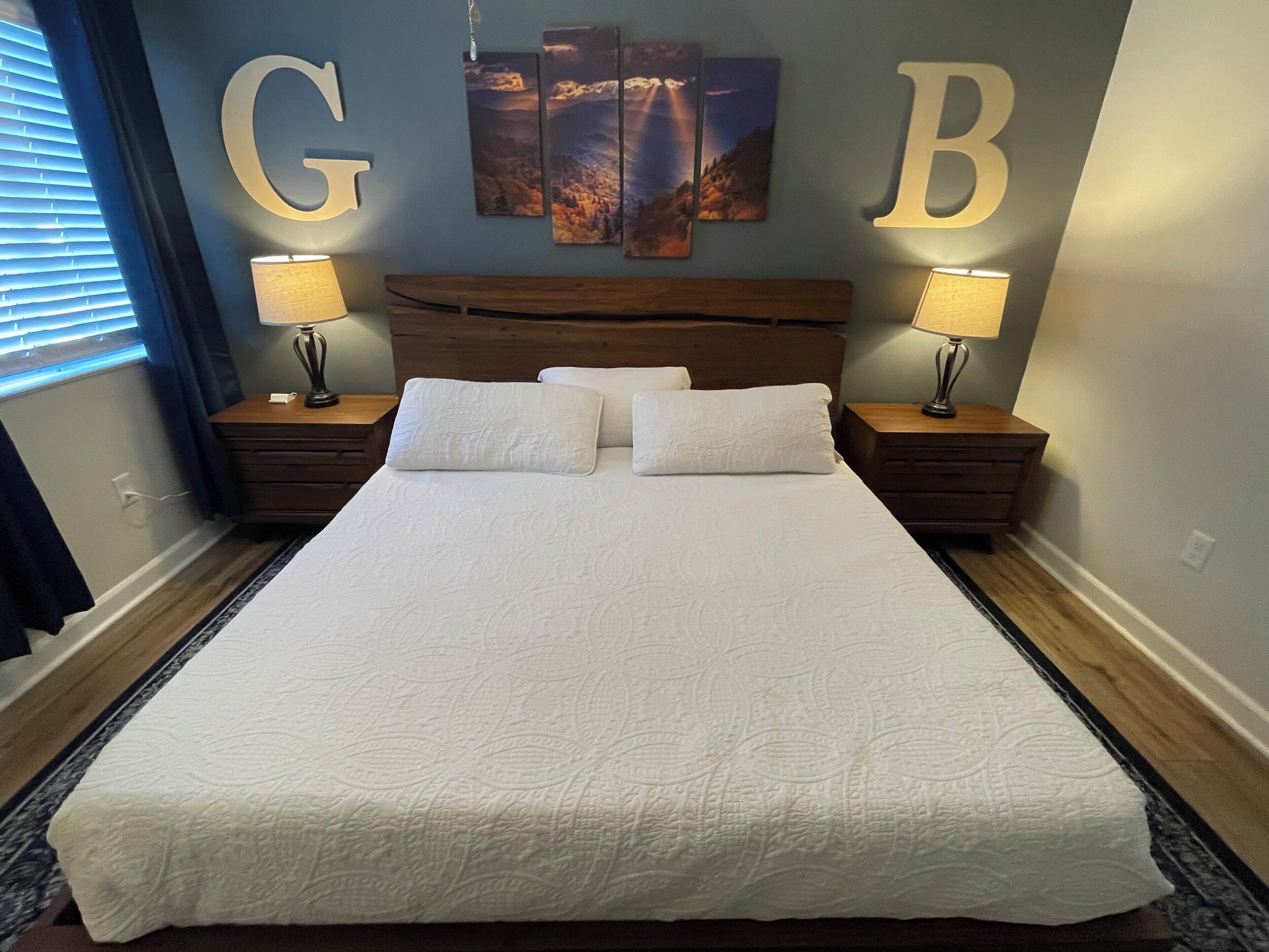 Een net opgemaakt bed met een hoofdbord, twee nachtkastjes met lampen, en wanddecoratie met de letters 'G' en 'B'.
