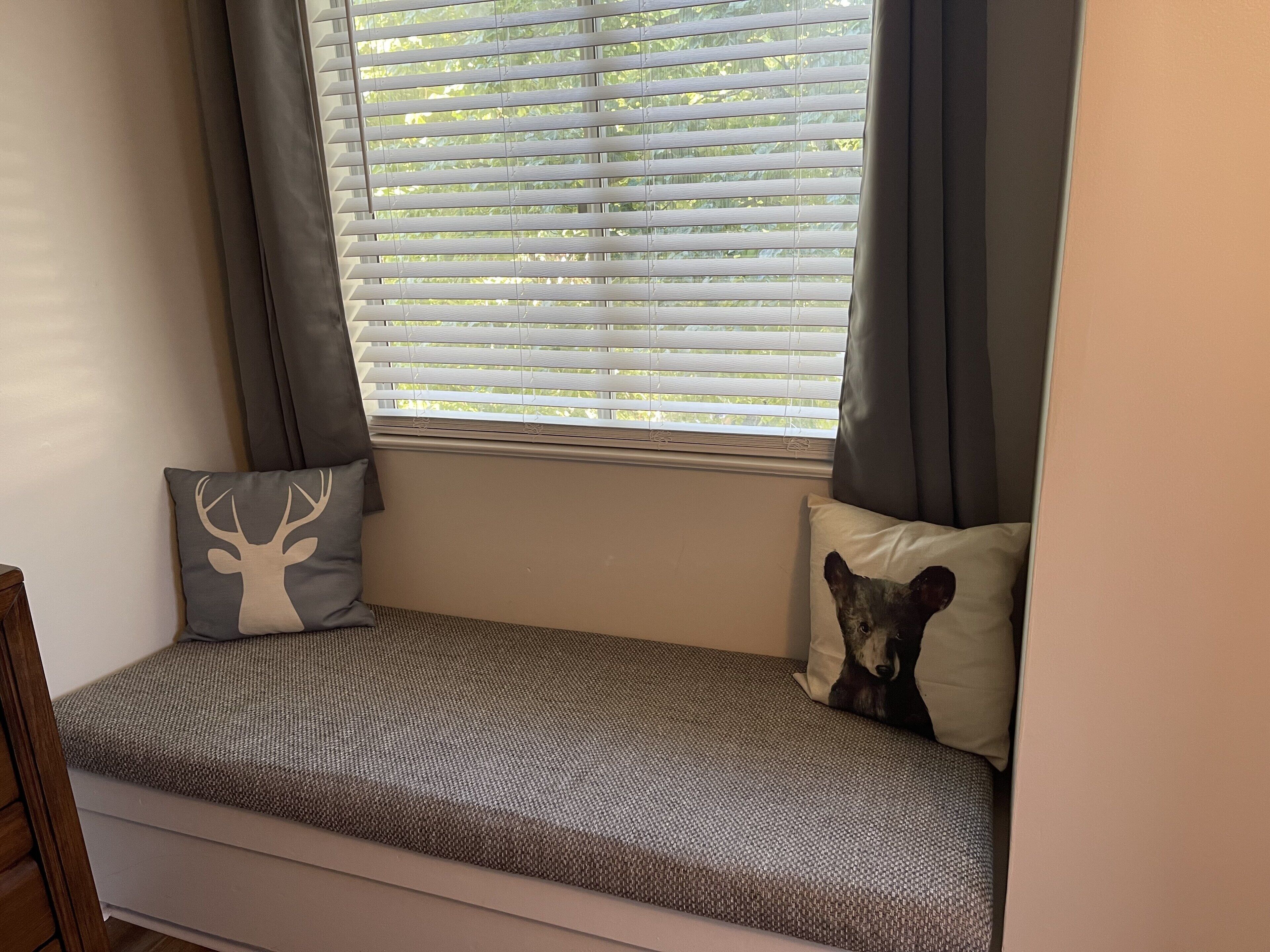 Un siège de fenêtre avec un banc en osier, deux coussins décoratifs et une vue sur la verdure à travers des stores blancs.