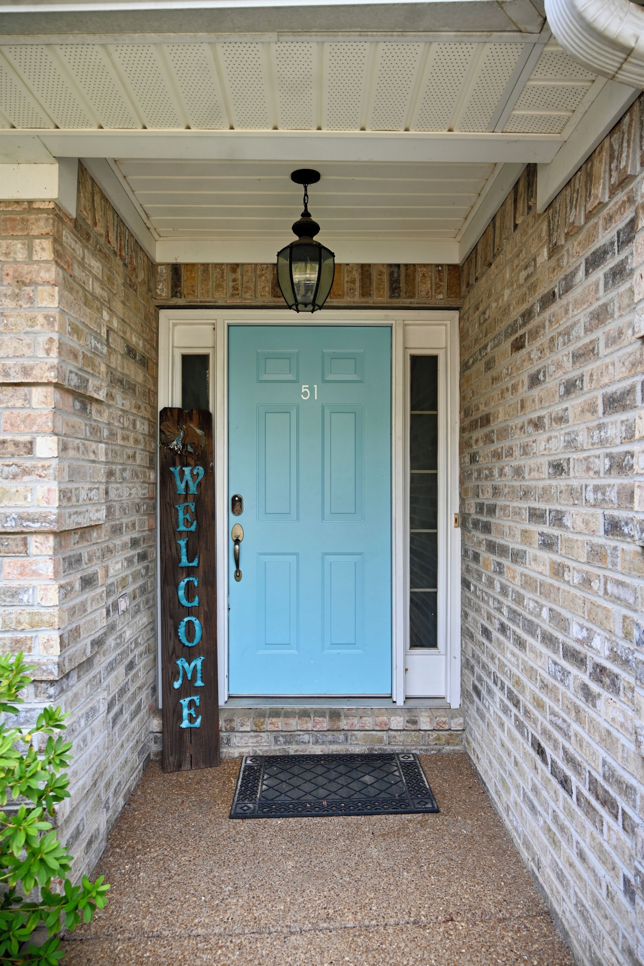 A blue door with a white doorbell, a welcome sign, and a black doormat on a brick wall.