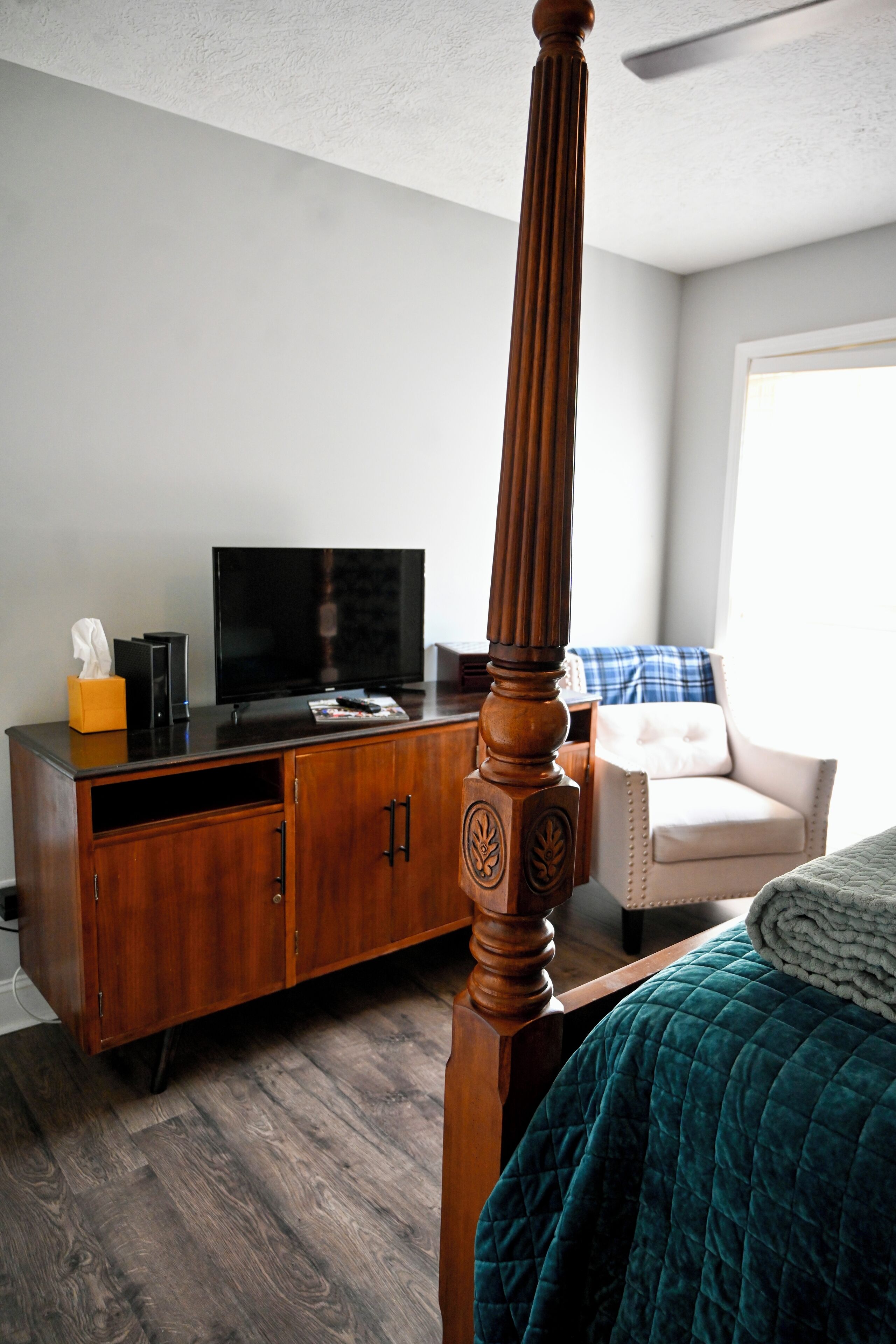 A bedroom with a wooden four-poster bed, a television on a wooden cabinet, and a chair with a blue and white blanket.