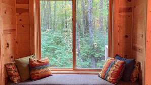 Una cabaña de madera con un banco incorporado bajo un ventanal grande, con vistas a un bosque.