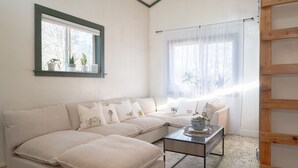 Ein Wohnzimmer mit einem beigefarbenen Ecksofa, einem Glastisch und einem Fenster mit transparenten Vorhängen.
